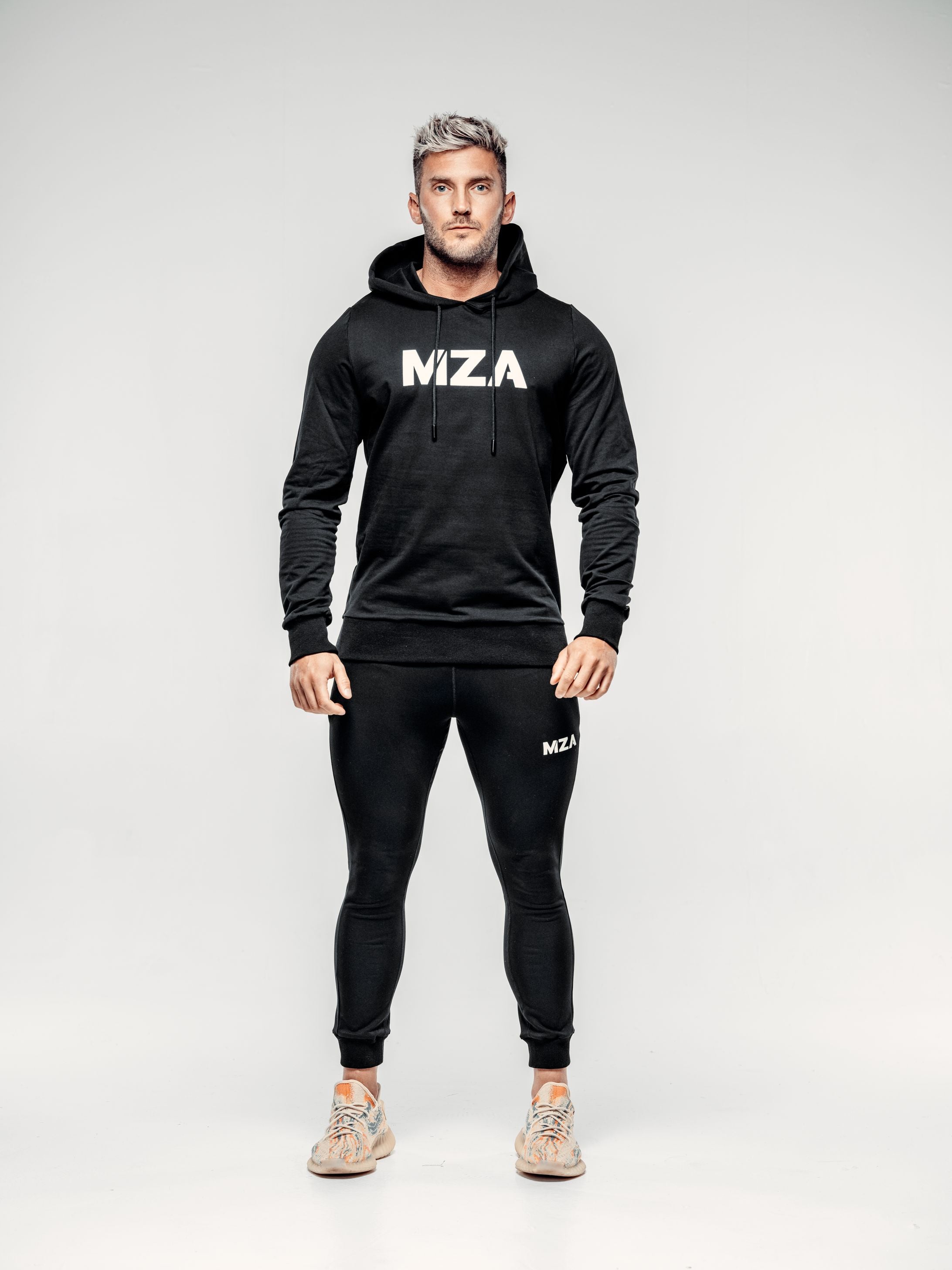 This is a full body shot of Shane straight on wearing the new standard hoodie in black.  This features the MZA logo in white in the middle of his chest.  Shane is also wearing the matching new standard joggers in black.
