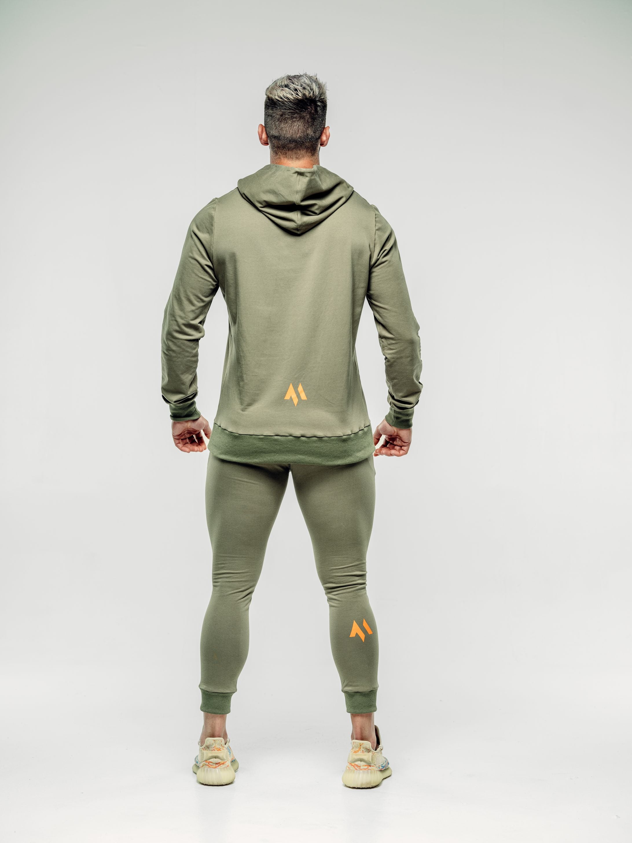 This is a rear view full body shot of Shane wearing the new standard hoodie in khaki.  It shows the M emblem in signature orange in the lower middle of the back as well as on the back of Shane's calf.