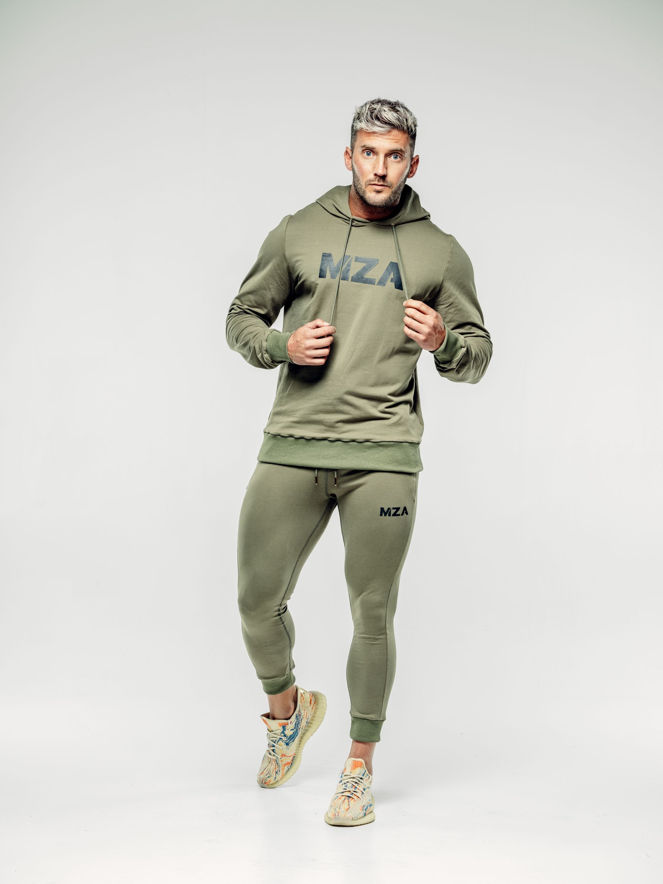 This is Shane wearing the New standard hoodie and new standard joggers 2 piece in khaki.  Shane is taking a step forward and is holding both strings from the neck of his hoodie
