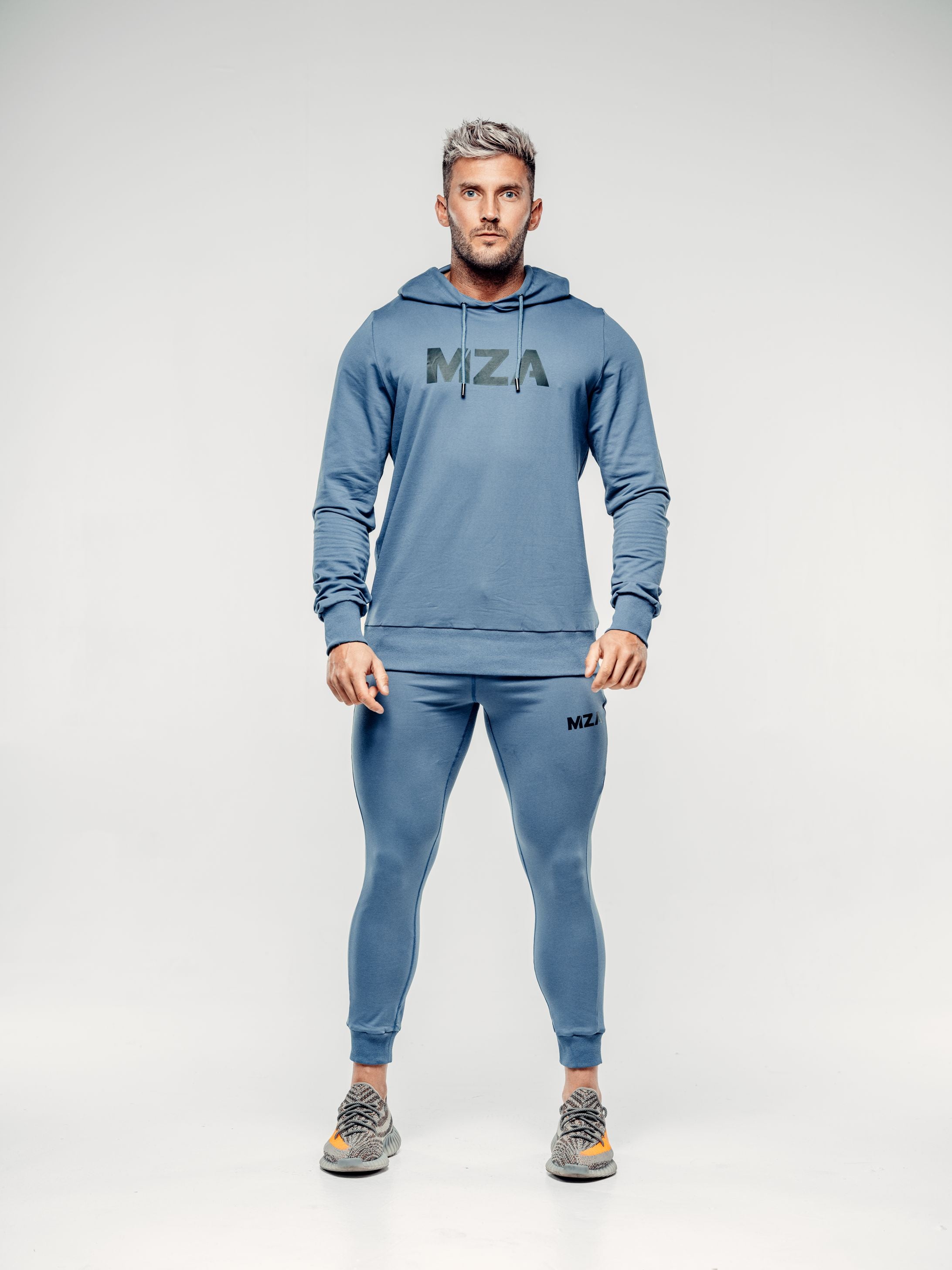 This is a full body shot of Shane straight on wearing the new standard hoodie in blue steel. This features the MZA logo in white in the middle of his chest. Shane is also wearing the matching new standard joggers in blue steel.