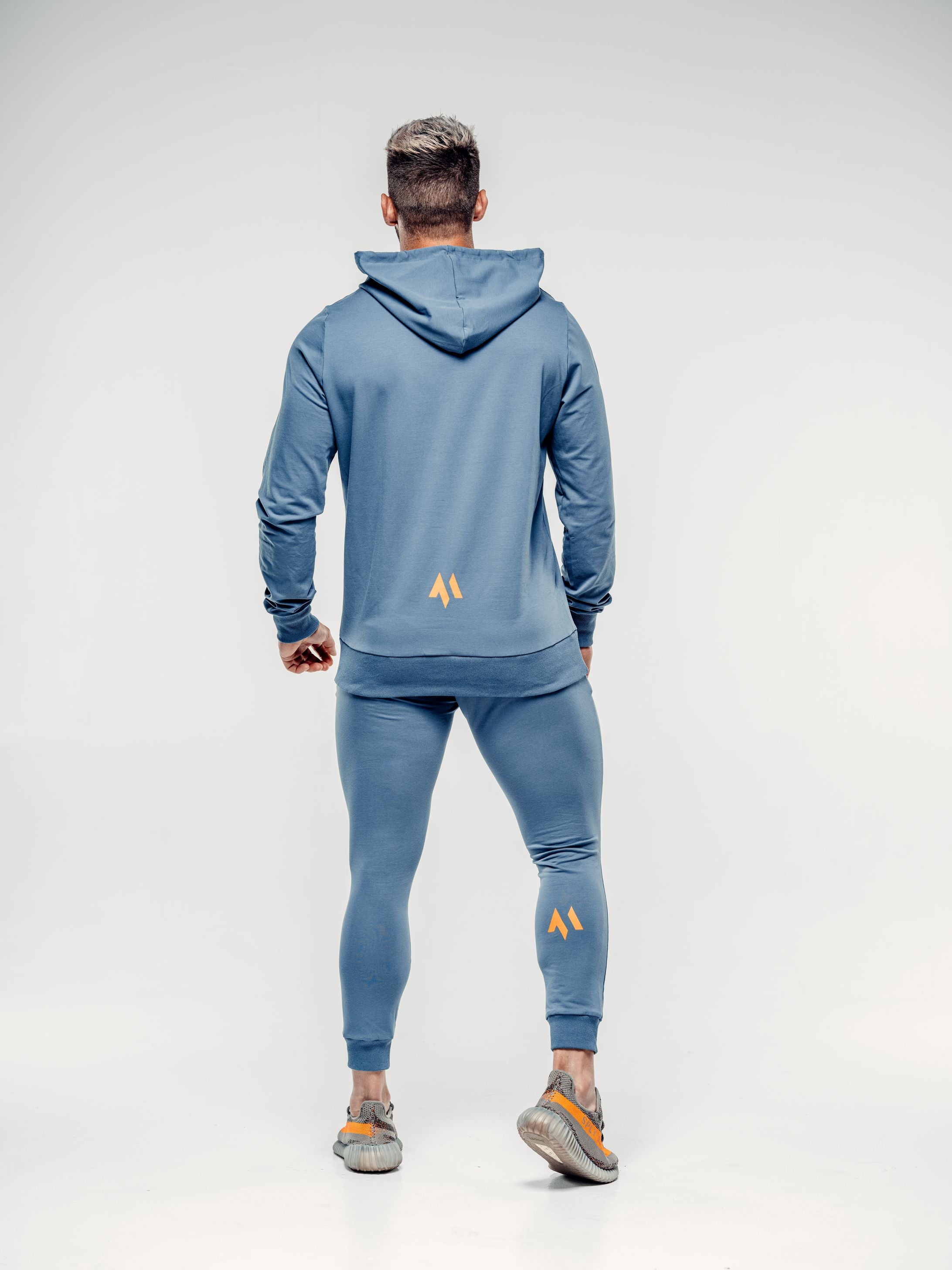 This is a rear view full body shot of Shane wearing the new standard hoodie in blue steel.  It shows the M emblem in signature orange in the lower middle of the back as well as on the back of Shane's calf.