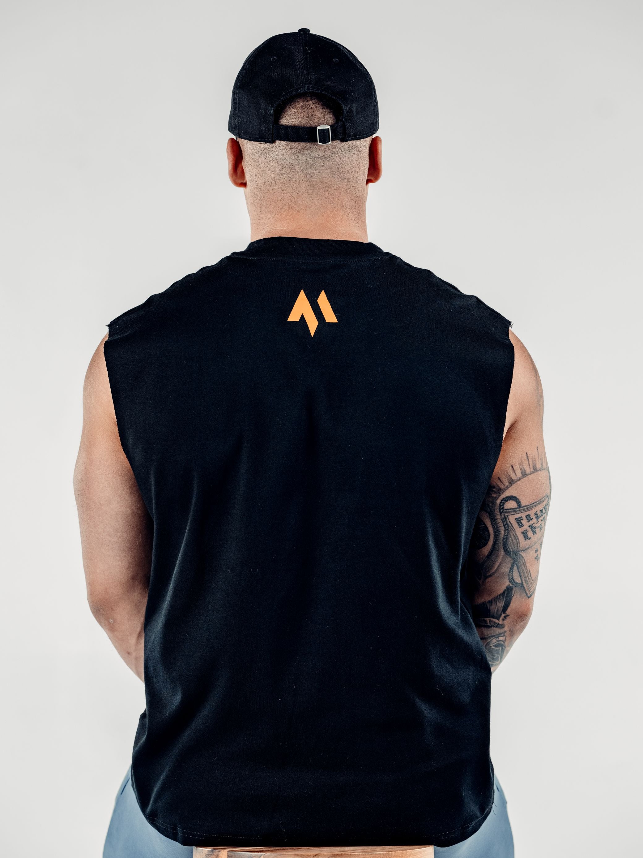 This is a rear view of Lewis, showcasing the back of the new standard 3d distressed 5 panel in black.  It showcases the strap and the buckle.  Lewis is also wearing the new standard vest in black which features the orange M emblem at the top of his back