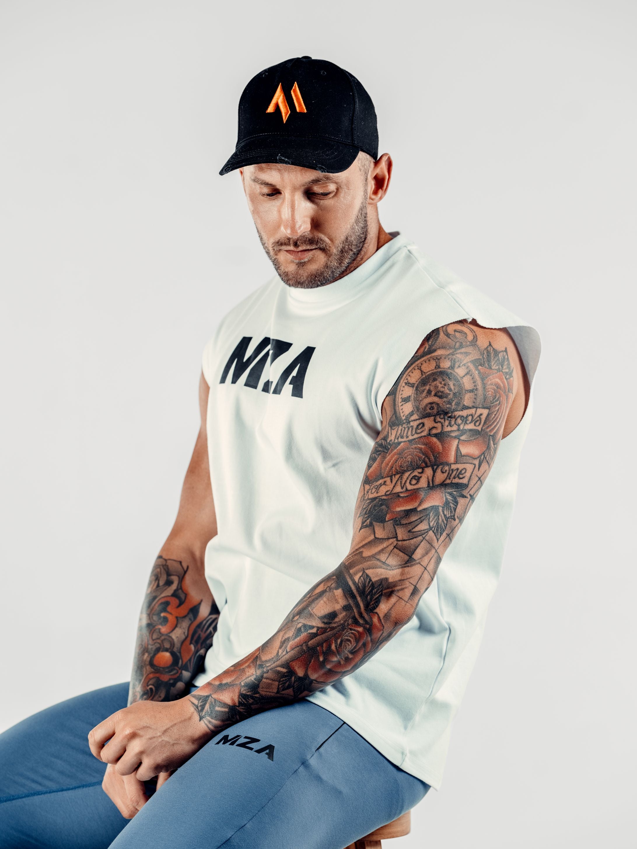 This is Shane sat on a stool facing the camera wearing the black new standard 3d distressed 5 panel black with an orange emblem.  He is wearing the new standard vest in white and the new standard joggers in blue