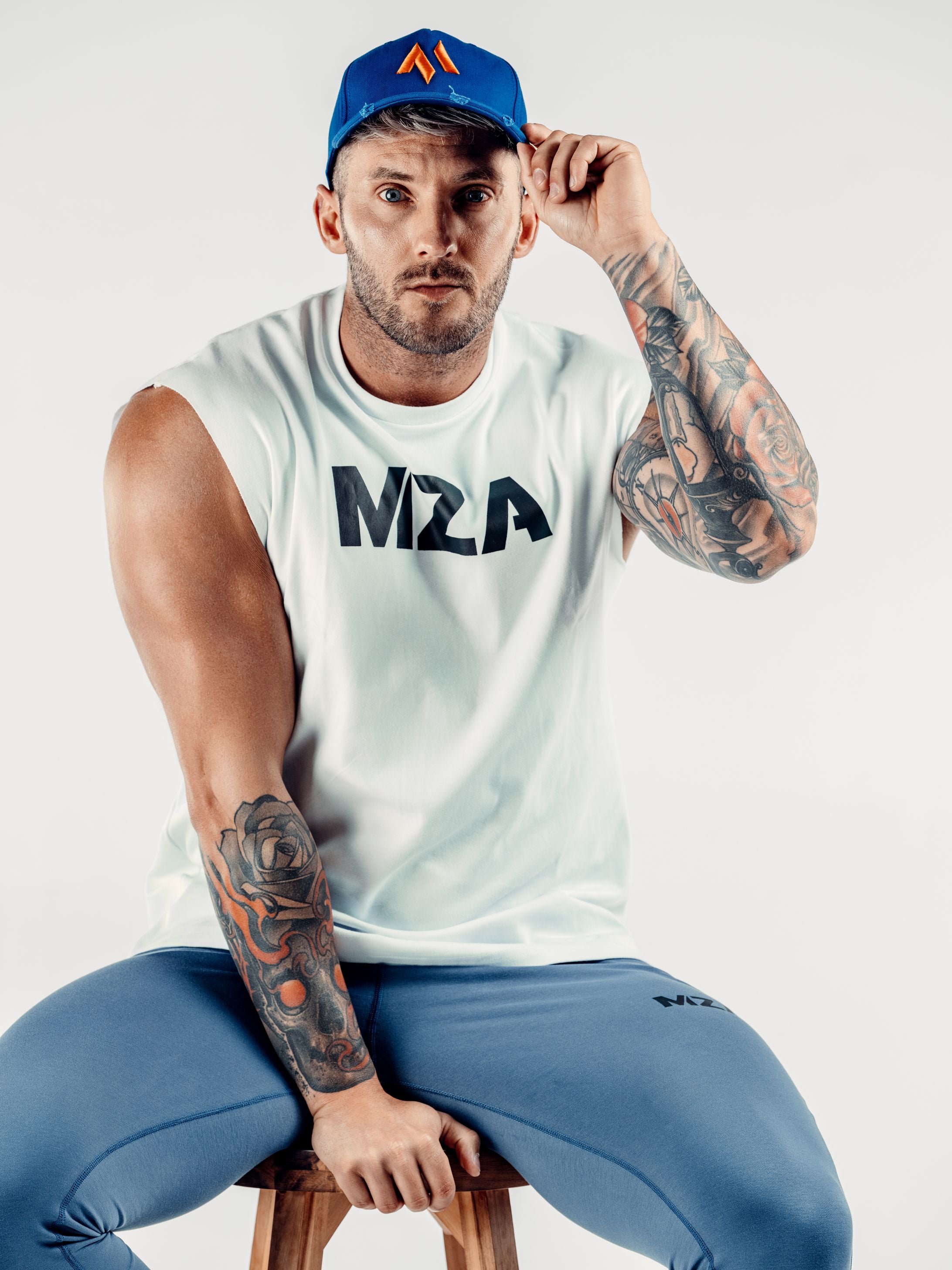 This is Shane wearing the new standard 3d distressed 5 panel in electric blue. It features the M emblem in orange. Shane is holding on to the peak with his right hand. Shane is also wearing the new standard vest in white which features the MZA logo on his chest and the new standard joggers in blue.