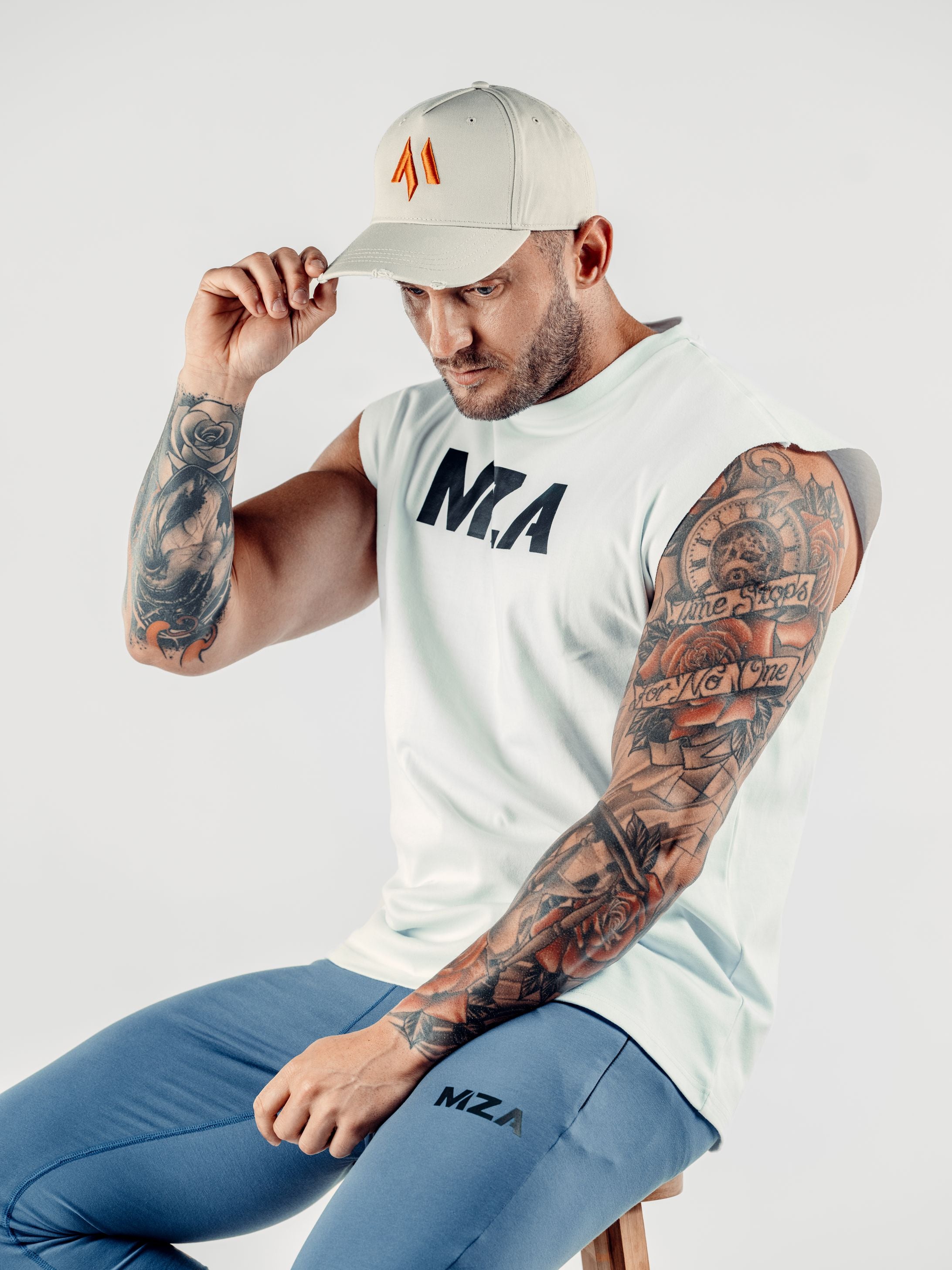 This is Shane wearing the new standard 3d distressed 5 panel in stone.  He is sitting on a slight angle with his right hand gripping the peak of the hat looking slightly down.  Shane is also wearing the new standard vest in white and the new standard joggers in blue.
